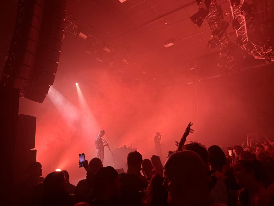 Pöbel MC auf der Bühne des FZW Dortmund. Die Lichtshow taucht die Halle in rotes Licht, im Vordergrund ist das Publikum zu sehen.